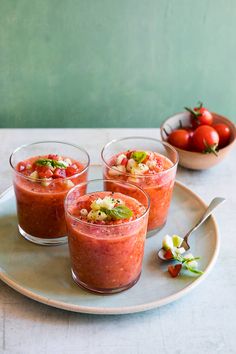 three small glasses filled with food on top of a white plate next to a bowl of tomatoes