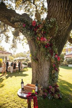 a table with wine bottles and flowers on it under a tree at a wedding reception