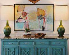 a painting hangs on the wall above a blue cabinet with two lamps and vases