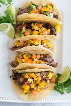 three tacos on a white plate with limes and cilantro