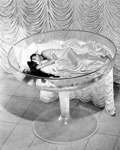 black and white photograph of a woman laying in a glass bowl on the floor next to a curtain