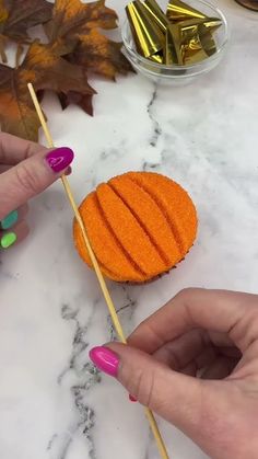 two hands holding sticks with an orange cake on it and some leaves in the background