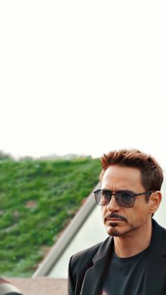 a man wearing sunglasses and a black suit looks off into the distance as he stands in front of a grassy area