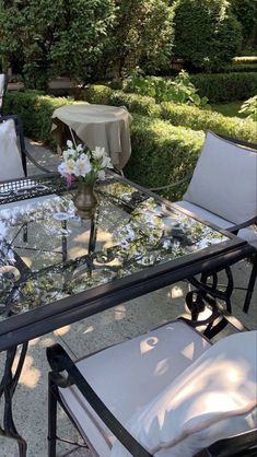 a glass table with white chairs around it in the middle of some bushes and trees