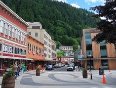 a street with shops and people walking on the sidewalk in front of some trees, mountains and buildings