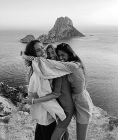 three young women hugging each other on top of a hill next to the ocean with an island in the background