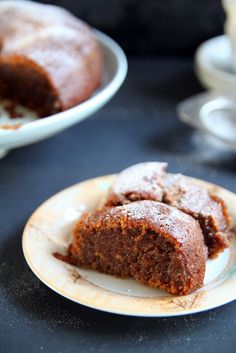 a piece of chocolate cake on a plate with another slice in the background and coffee cups to the side