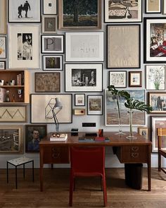 a wall full of framed photographs and pictures with a red chair in front of it