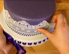 a woman is decorating a cake with white lace and purple ribbon on it's edge