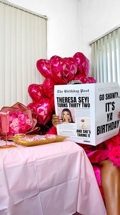 a woman sitting at a table reading a newspaper with pink balloons on top of it