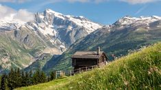 an old cabin in the mountains with snow on the mountain tops behind it and green grass below