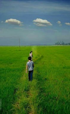 two people walking in the middle of a green field with blue sky and white clouds