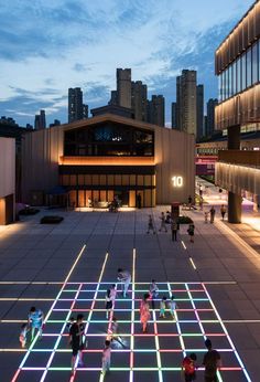 people are walking around in an open area with lights on the floor and buildings behind them