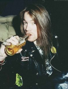 a man with long hair is drinking from a beer glass in his hand and wearing a leather jacket