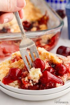 a person holding a fork over a piece of pie with cherries on it in a white bowl