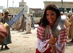 a woman is holding two large knives and smiling at the camera while other people stand around her