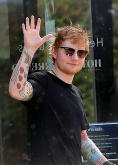 a man with red hair and tattoos waves to the camera while standing in front of a building