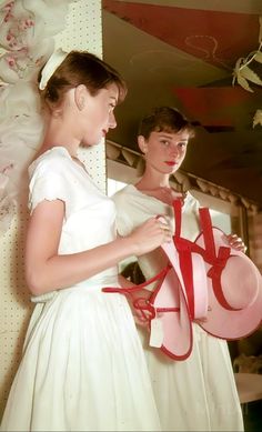 two women in white dresses are holding red ribbons and hats while standing next to each other