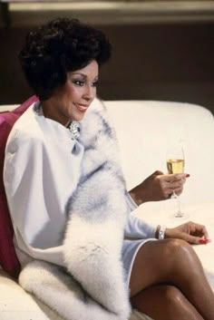 a woman sitting on a couch with a glass of wine in her hand and wearing a fur coat