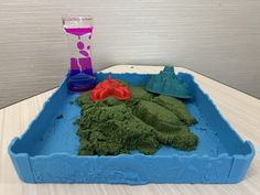 a blue tray filled with green sand next to a purple cup and plastic toy boat