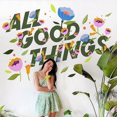 a woman standing in front of a wall with flowers on it and the words all good things above her