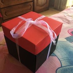 a red and black gift box with a white ribbon on the top sitting on a bed