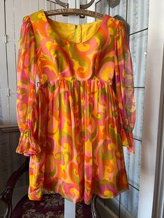 This fabulous dress by Joyce Palmer is made of medium weight golden yellow fabric with a sheer polyester print overlay in a large paisley type pattern, in shades of orange, yellow, green and light and dark pink. The sleeves are sheer with gathers at the cuff (please note that the elastic is no longer stretchy). The dress has darts at the bust and an empire waist, and fastens with a hook and eye and long zipper in the back. Marked size 7/8. The measurements, taken with the dress lying flat, are: 1970s Style Orange Summer Dress, Retro Orange Dresses With Vibrant Print, Retro Orange Dress With Vibrant Print, Vintage Orange Mini Dress For Spring, 1970s Style Mini Summer Dress, Green Print Dress, 1960s Dresses, Orange Floral Print, Fabulous Dress