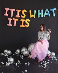 a woman sitting in front of balloons and confetti with the words it's what it is