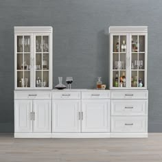 two white cabinets with wine glasses and bottles on them in front of a gray wall