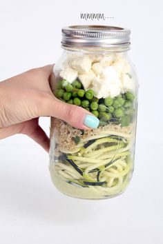 a hand holding a mason jar filled with pasta, peas and cucumber slices