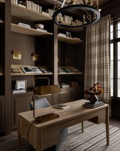 a desk with a lamp, bookshelf and flowers on it in front of a window