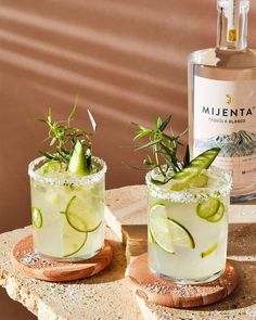 two margaritas with lime slices and garnish sit on a table next to a bottle