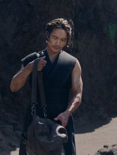 a man holding onto a bag while standing on top of a dirt field next to a mountain