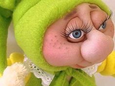 a close up of a stuffed animal wearing a green outfit with eyes wide open and long eyelashes