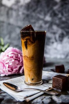 a tall glass filled with liquid next to a pink flower and some chocolate pieces on a table