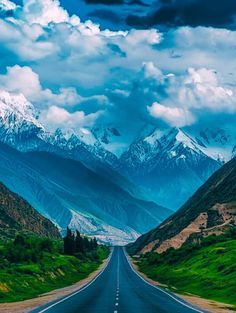 an empty road in the middle of mountains with snow capped peaks on each side and green grass below