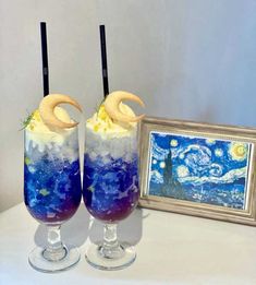 two glasses filled with blue and white liquid next to a painting on a table top