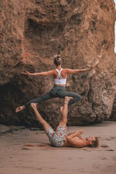 two people doing acrobatic tricks on the beach near some large rocks in front of them