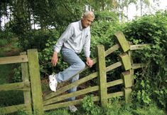 an older man jumping over a wooden fence in the grass and bushes, with his foot on the gate