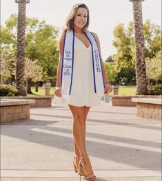 a woman in a white dress and blue sash is posing for the camera with palm trees behind her