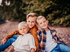 three children sitting on the ground in front of some trees and bushes with their arms around each other