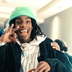 a young man with dreadlocks and a green beanie smiles as he holds his hand up to his ear