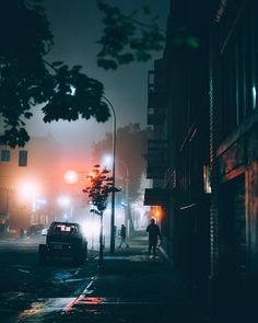 a person walking down the street at night in the foggy city with cars parked on the side walk