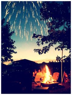 people sitting around a fire pit with fireworks in the sky above them and trees on either side