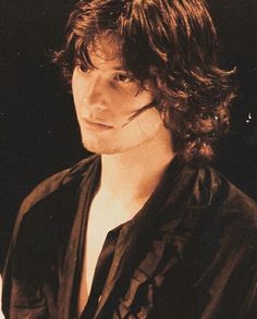 a young man with long hair wearing a black shirt and looking at the camera while standing in front of a dark background