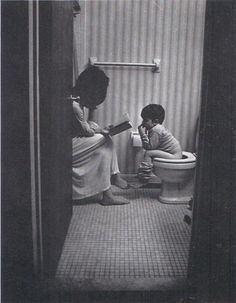 two women are sitting on the toilet in a small bathroom, one reading a book