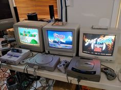 three video game consoles sitting on top of a table next to each other with two monitors