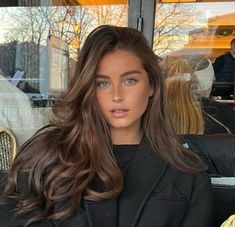 a woman with long brown hair sitting in front of a window and looking at the camera