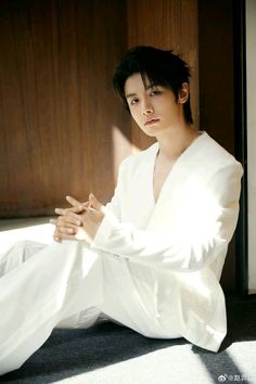 a young man sitting on the floor in white clothing with his hands folded out and looking at the camera