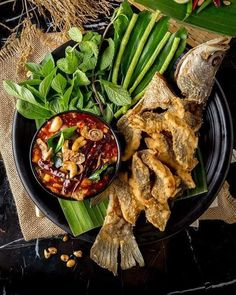 a black plate topped with fried fish and veggies next to a bowl of sauce